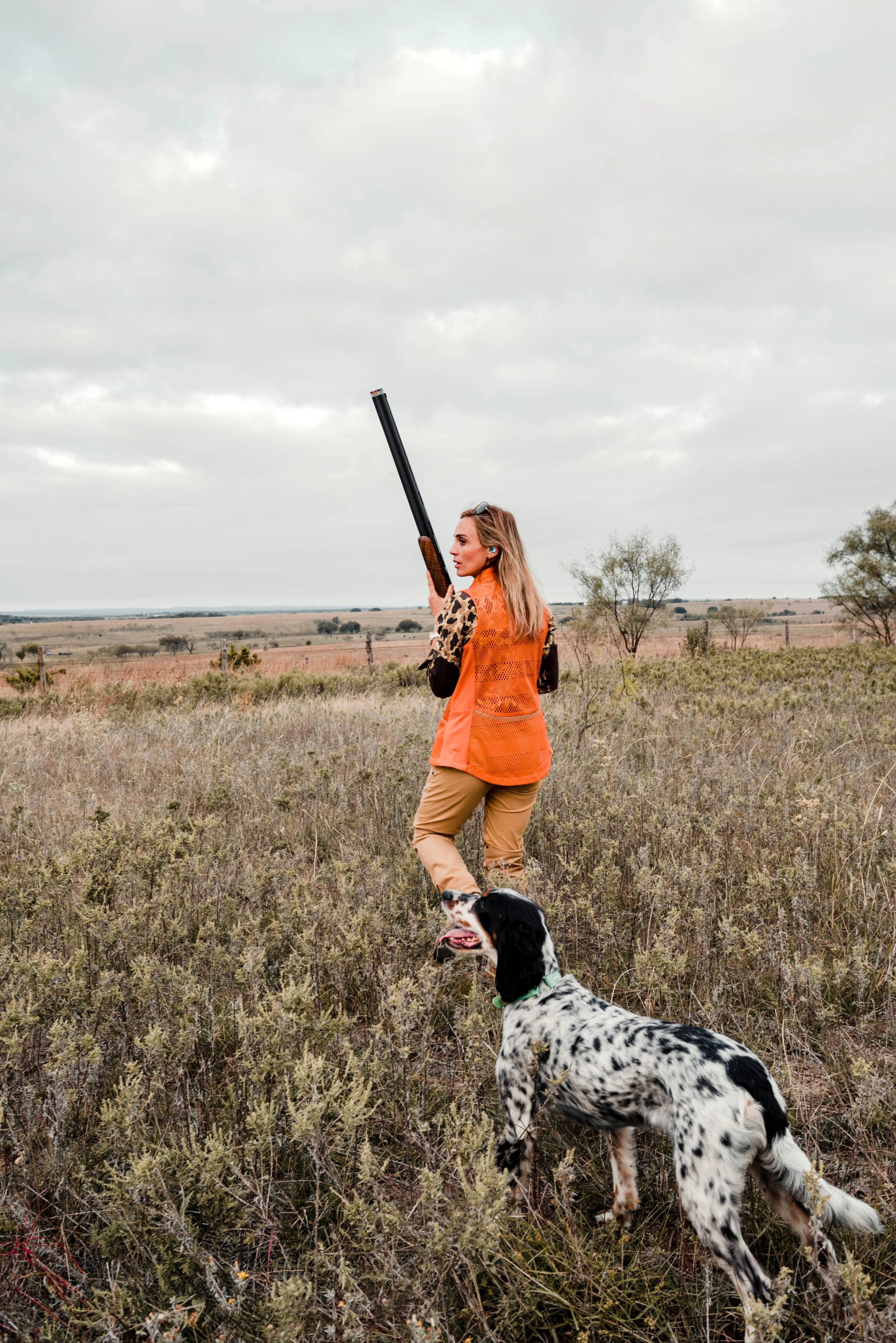 Woman in orange vest holding a shotgun with a dog in a field