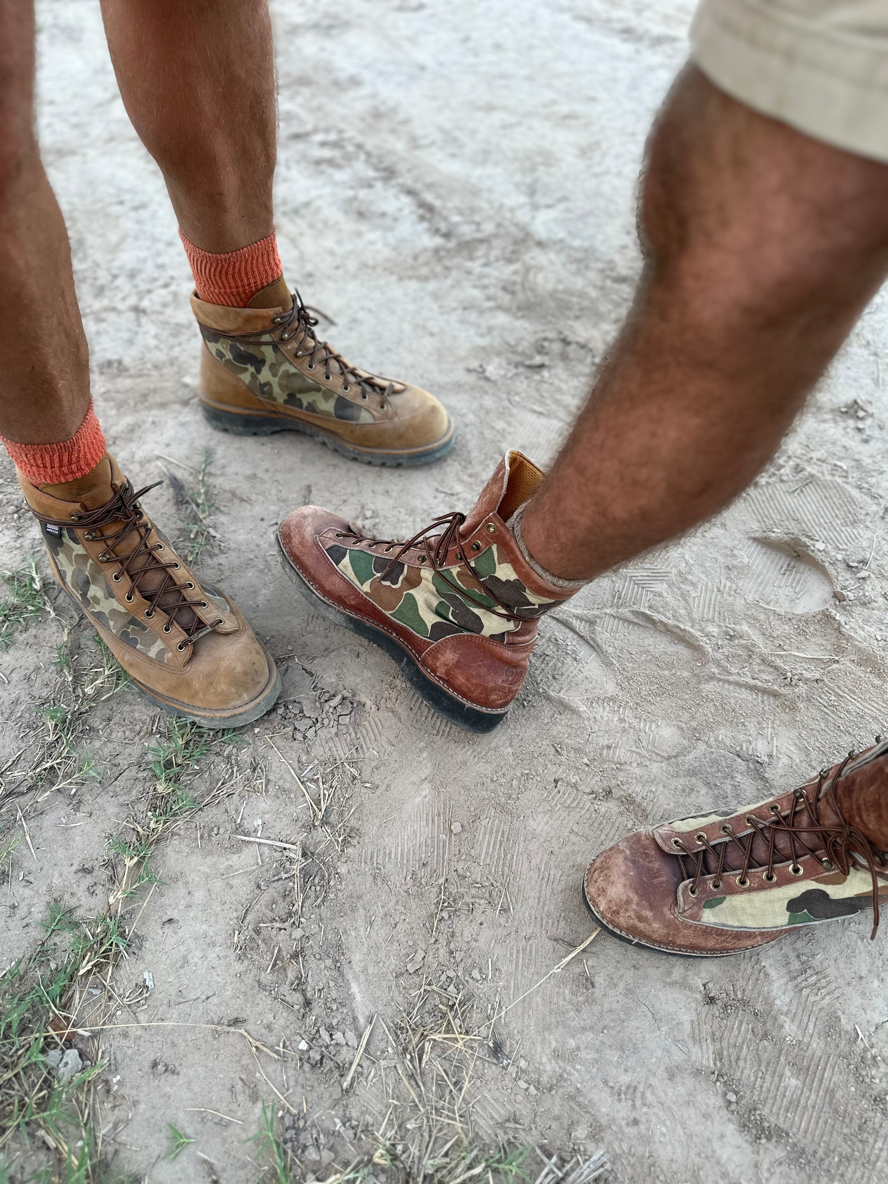 Two pairs of feet wearing camouflage boots on a sandy ground.