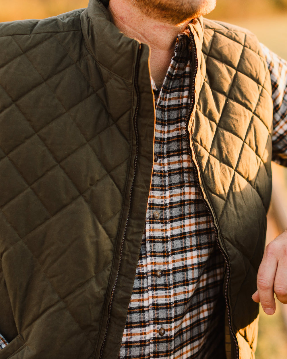 Man wearing a quilted vest over a plaid shirt with a blurred natural background
