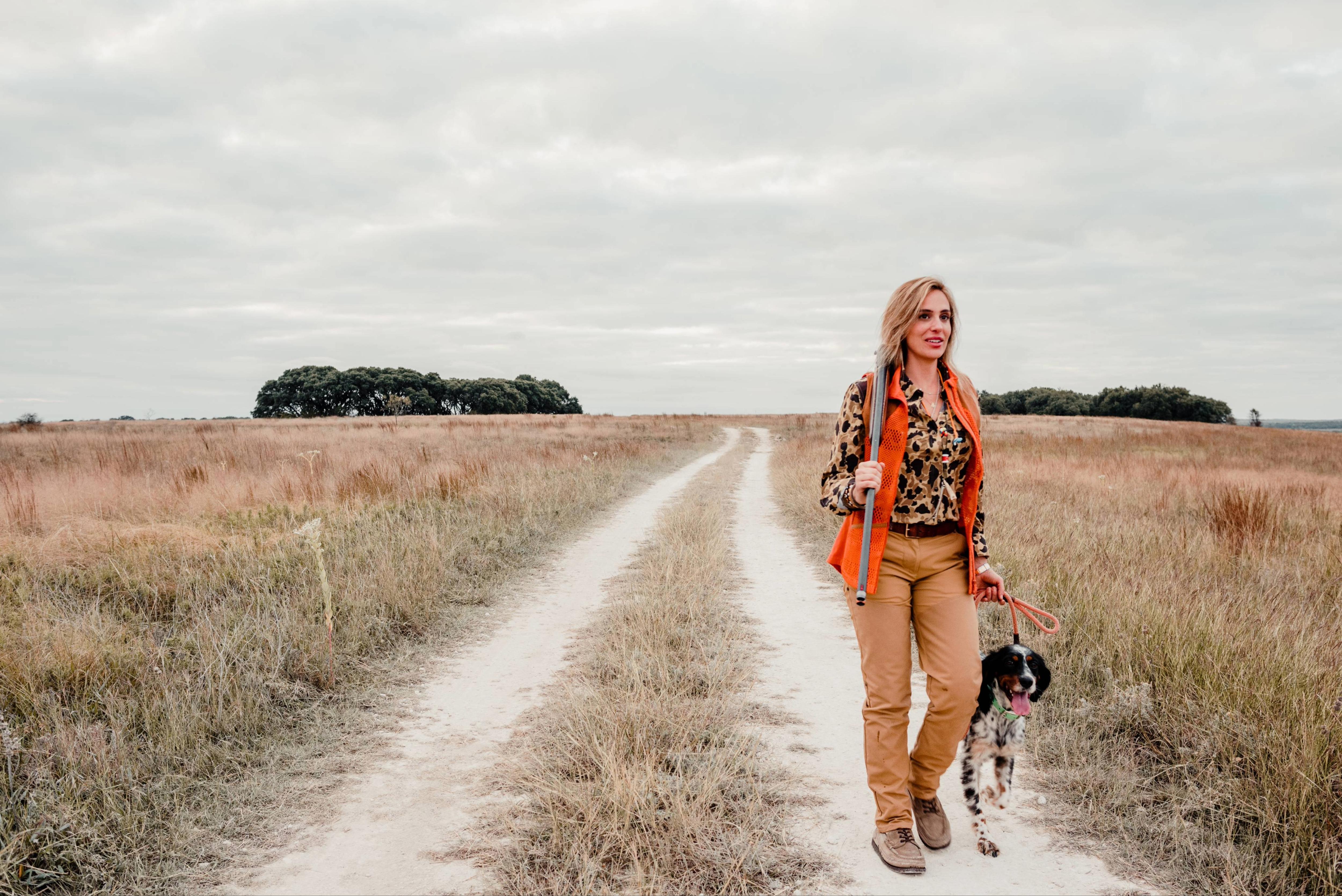 Woman walking a dog on a dirt path through a field with an orange jacket and patterned shirt.