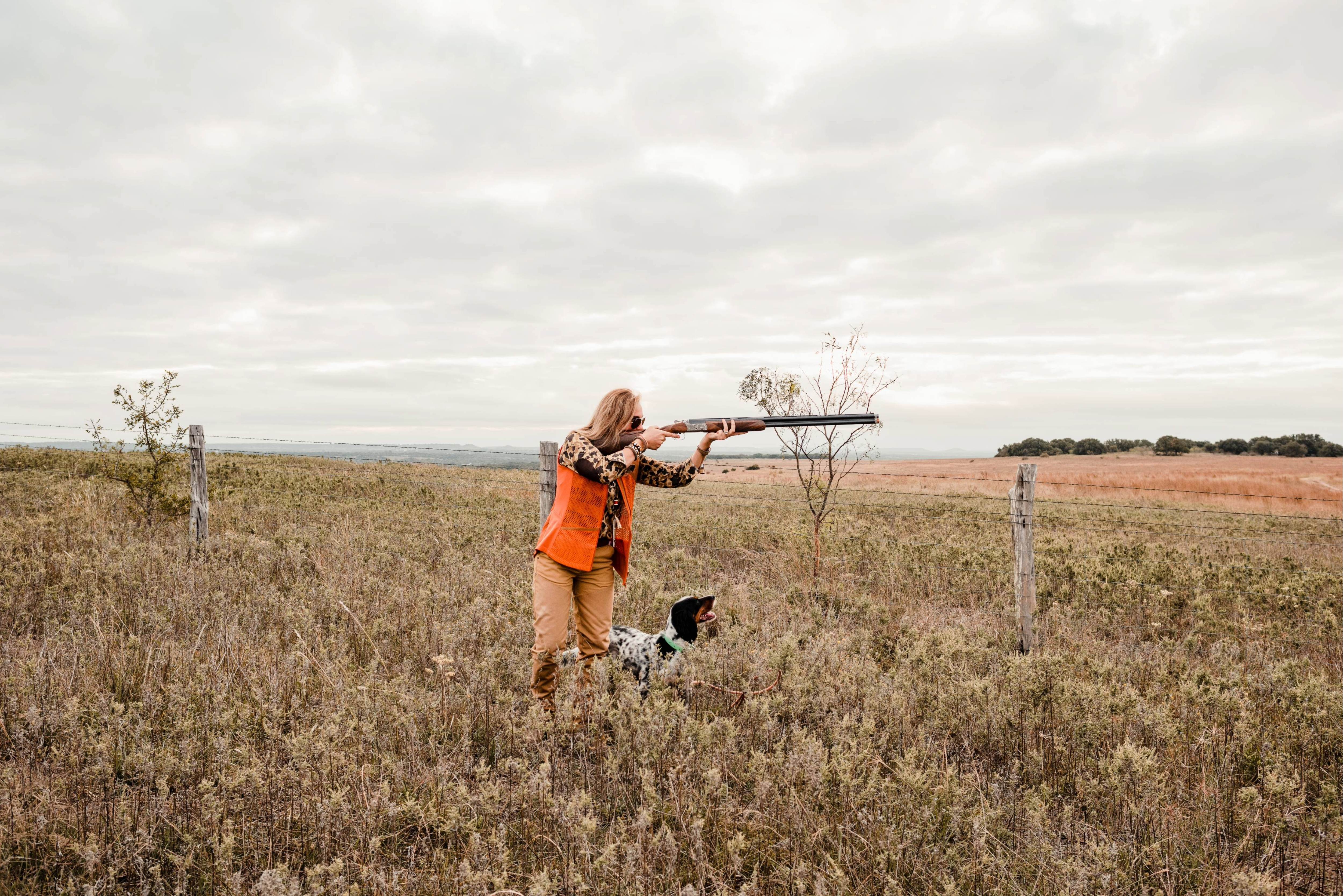 Person in orange hunting vest holding a shotgun with a dog in a field on a cloudy day