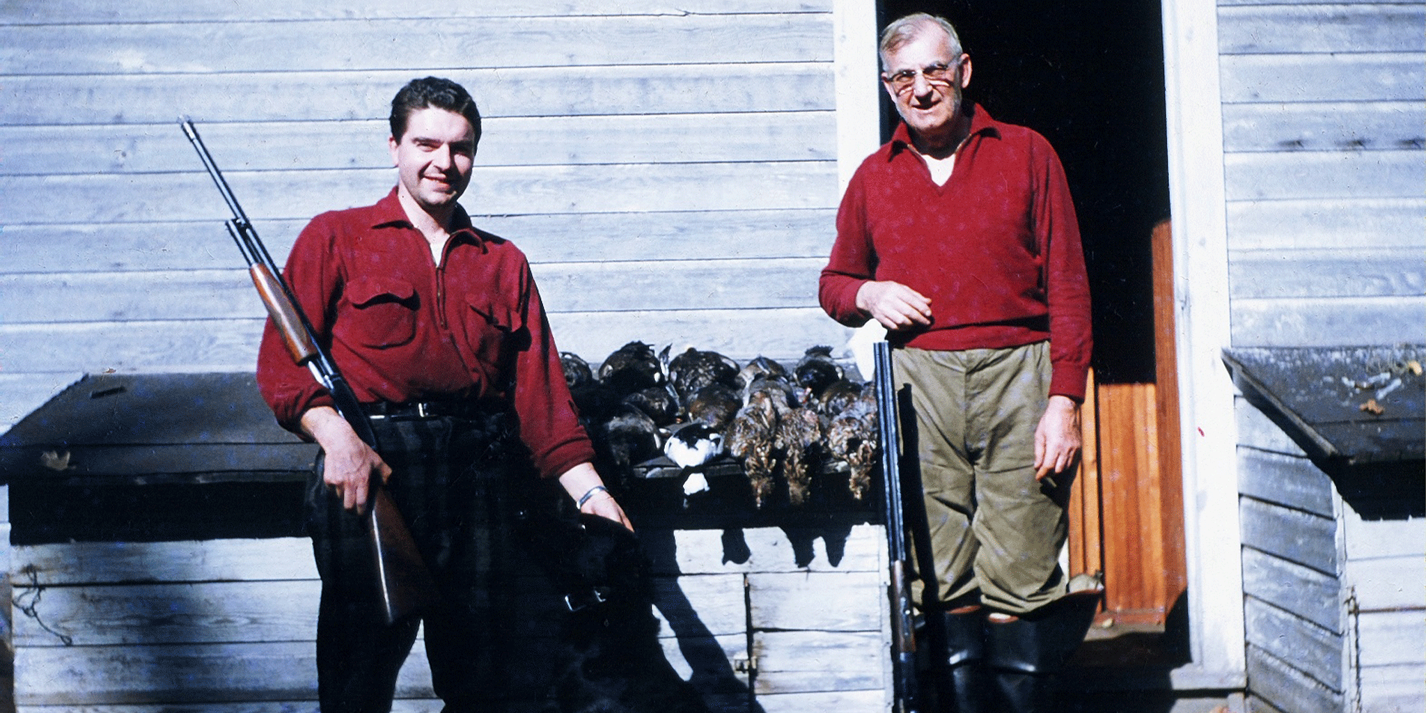 Two men in red shirts standing in front of a wooden shed with hunting equipment.